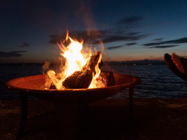 Beach fire pit with sandal wearing feet at sunset.