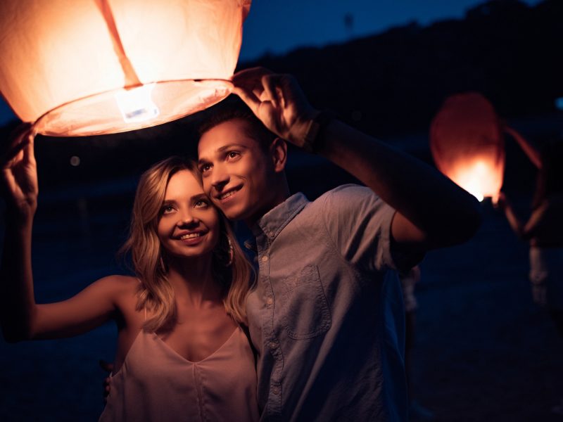young couple launching sky lantern on river beach in evening